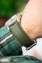 Close-up of a green textured watch strap with visible stitching, worn on a wrist and attached to a brushed metal case. The macro angle highlights the fabric-like texture and stitching details, while a green plaid sleeve and softly blurred background add depth and contrast.