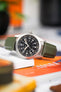 Close-up of a Hamilton-style field watch with a black dial and full Arabic numerals, resting on stacked books. The watch features a brushed metal case and is fitted with a green textured strap, while a blurred background of orange and dark tones adds contrast and depth.