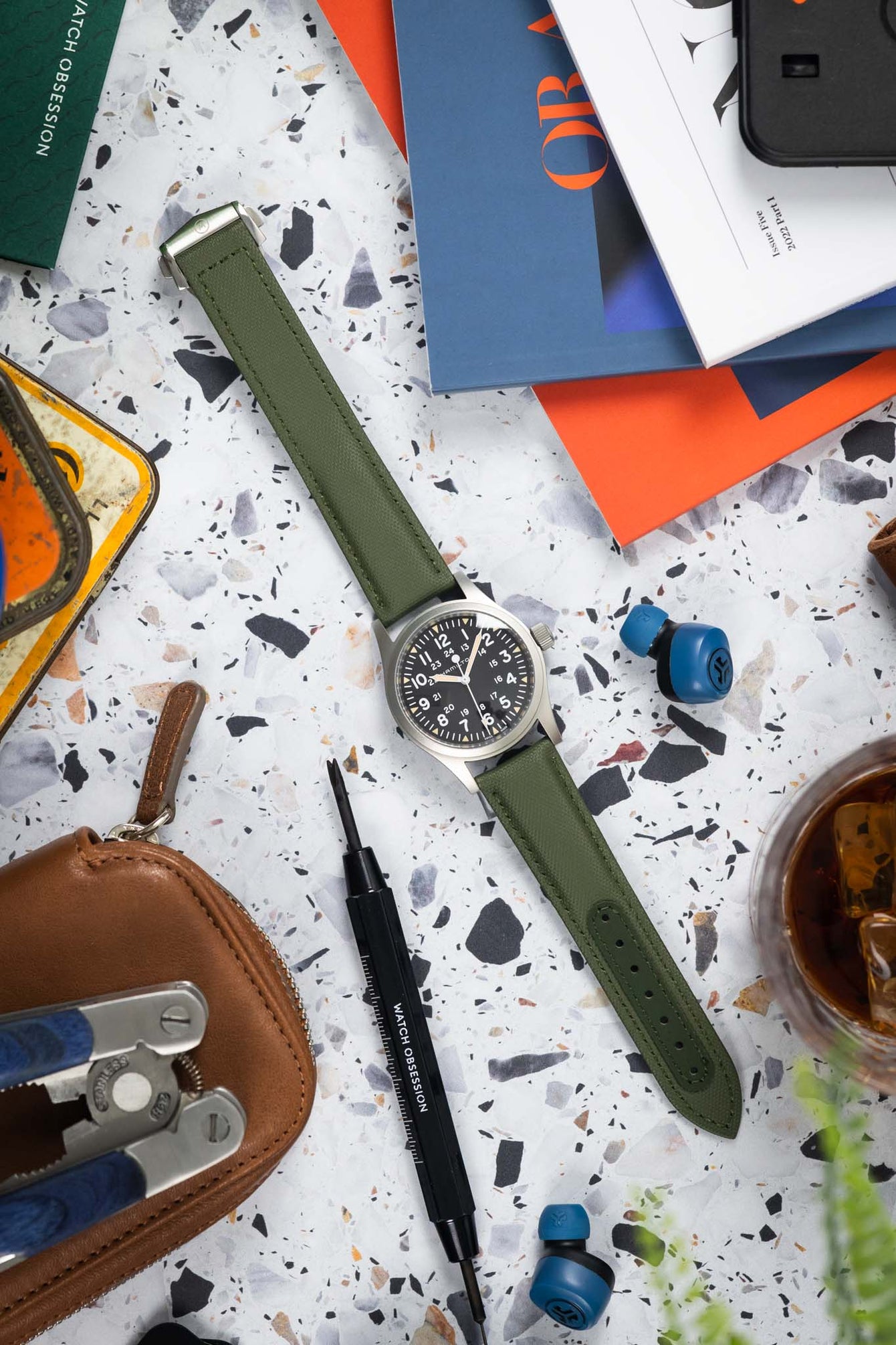 Flat lay of a Hamilton-style field watch with a black dial on a green textured strap, arranged on a speckled surface. The scene includes watch tools, a leather pouch, a vintage tin, magazines, earbuds, and a glass with a drink, creating a workshop-style composition with green, orange, and neutral tones.