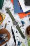 Flat lay of a Hamilton-style field watch with a black dial on a green textured strap, arranged on a speckled surface. The scene includes watch tools, a leather pouch, a vintage tin, magazines, earbuds, and a glass with a drink, creating a workshop-style composition with green, orange, and neutral tones.