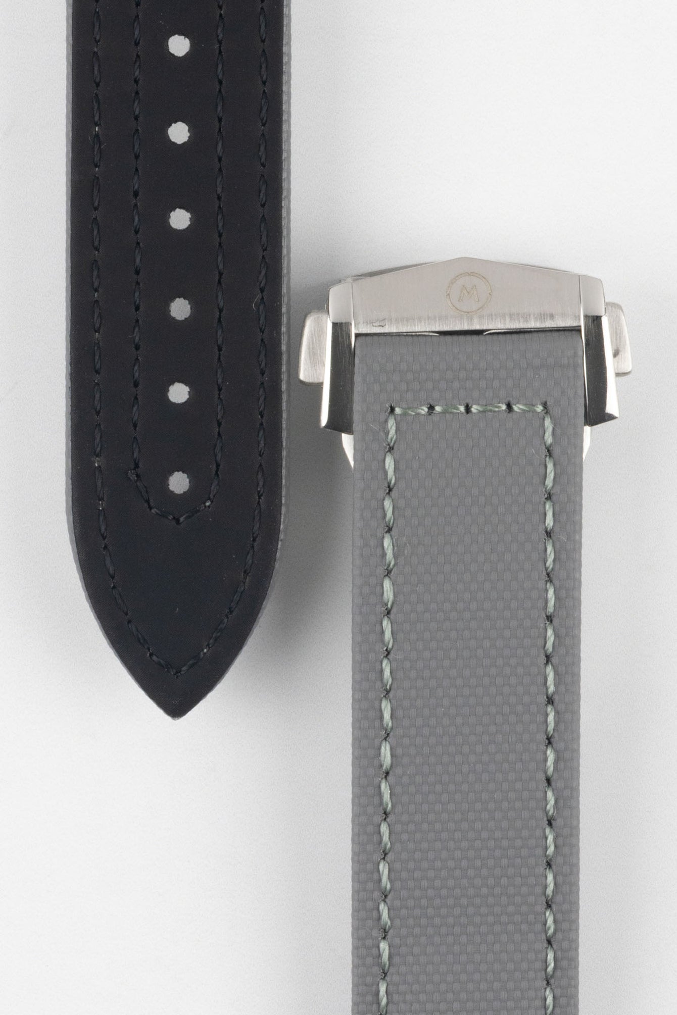 Two grey textured watch strap pieces displayed vertically on a light grey background, featuring subtle light grey stitching; one strap is fitted with a brushed silver deployant clasp with an engraved logo, while the other shows a row of evenly spaced adjustment holes down the center.