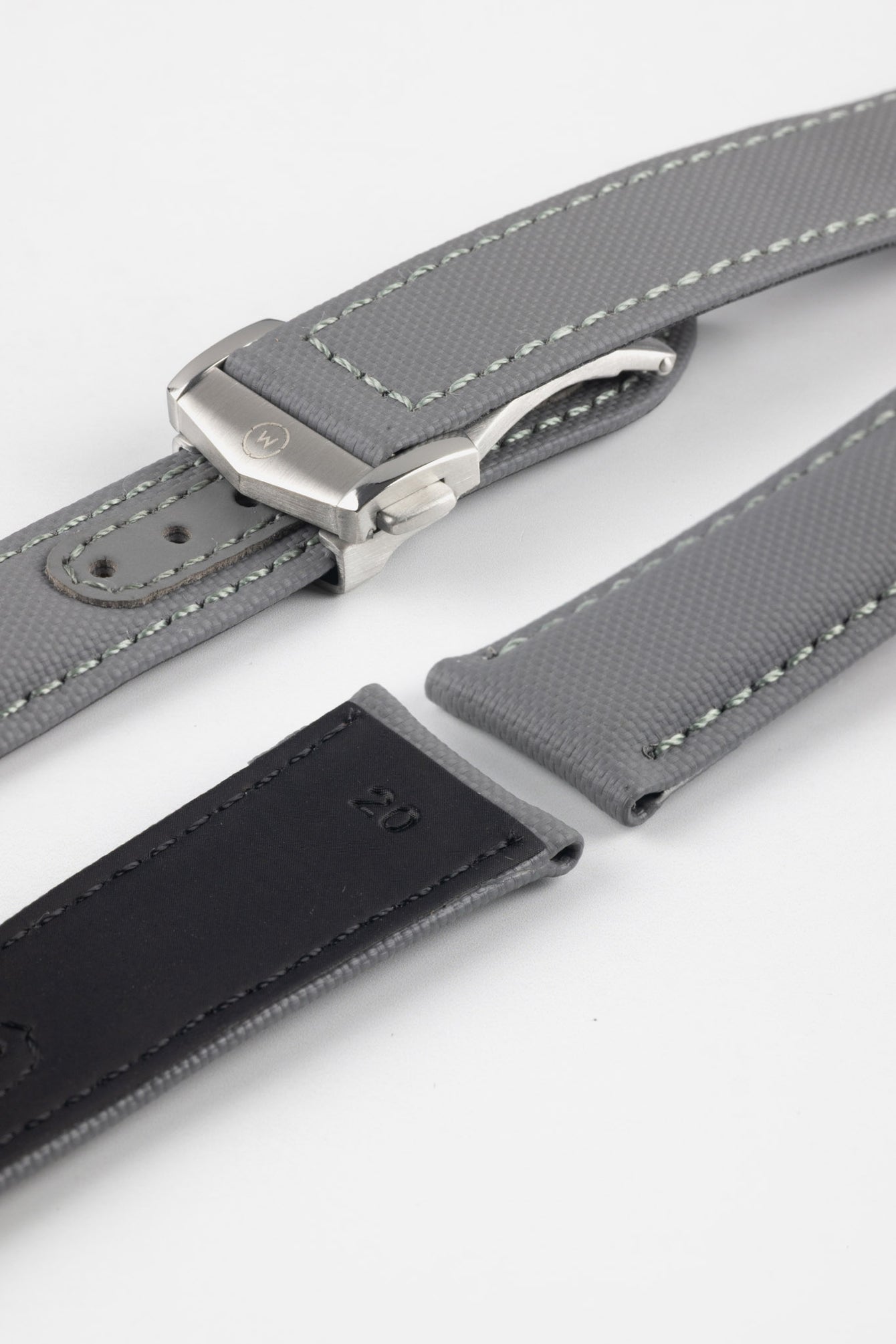 Angled close-up of a grey textured watch strap with light grey stitching, featuring a brushed silver deployant clasp with an engraved logo, alongside separate strap sections, all arranged on a light grey background.