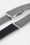 Angled close-up of a grey textured watch strap with light grey stitching, featuring a brushed silver deployant clasp with an engraved logo, alongside separate strap sections, all arranged on a light grey background.