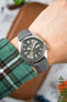 Close-up of a Seiko automatic watch with a grey sunburst dial and day-date display, worn on a wrist with a grey textured strap. The polished case and luminous markers stand out against a softly blurred background with warm tones and workshop elements.