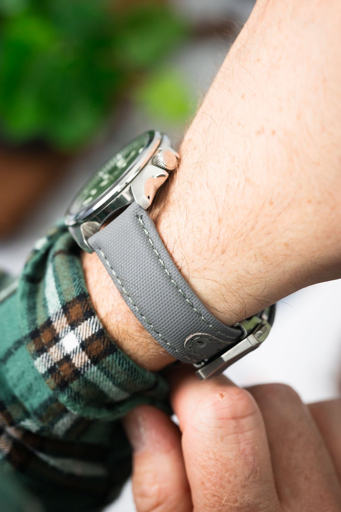 Close-up of a grey textured watch strap with stitching, worn on a wrist and attached to a polished metal case. The shallow depth of field highlights the strap’s fabric-like texture and stitching, with a soft, blurred background of green and neutral tones.