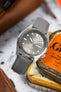 Close-up of a Seiko automatic watch with a grey sunburst dial and day-date display, resting on stacked vintage tins. The watch is fitted with a grey textured strap, and the polished case and luminous markers stand out against a softly blurred background with warm tones and workshop-style elements.
