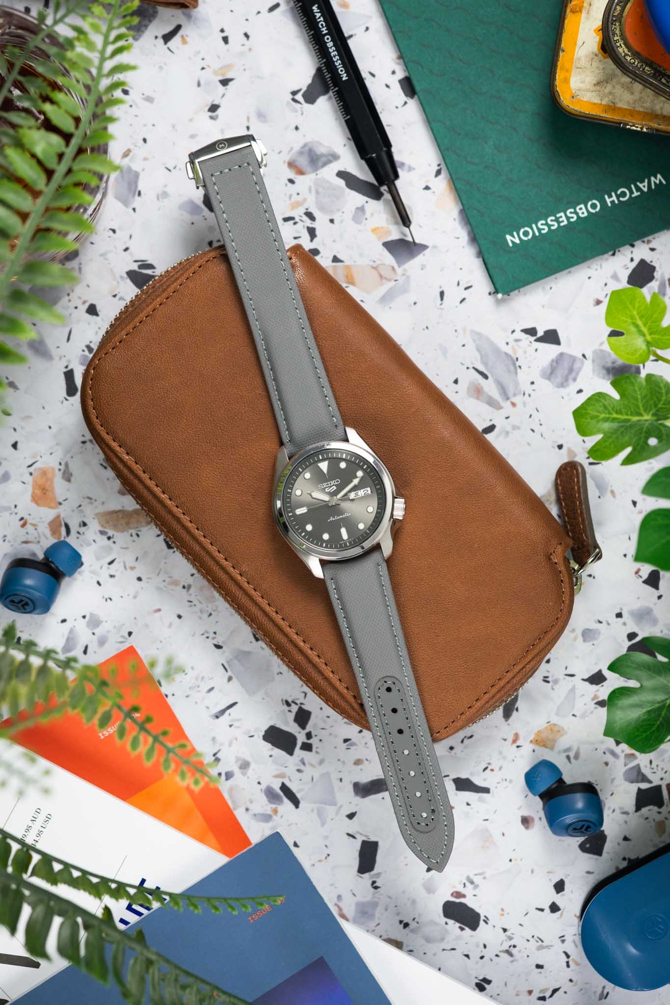 Flat lay of a Seiko automatic watch with a grey sunburst dial and day-date display on a grey textured strap, placed on a brown leather pouch. The scene is arranged on a terrazzo-style surface with watch tools, magazines, earbuds, greenery, and a vintage tin, creating a workshop-inspired composition with neutral, green, and orange accents.