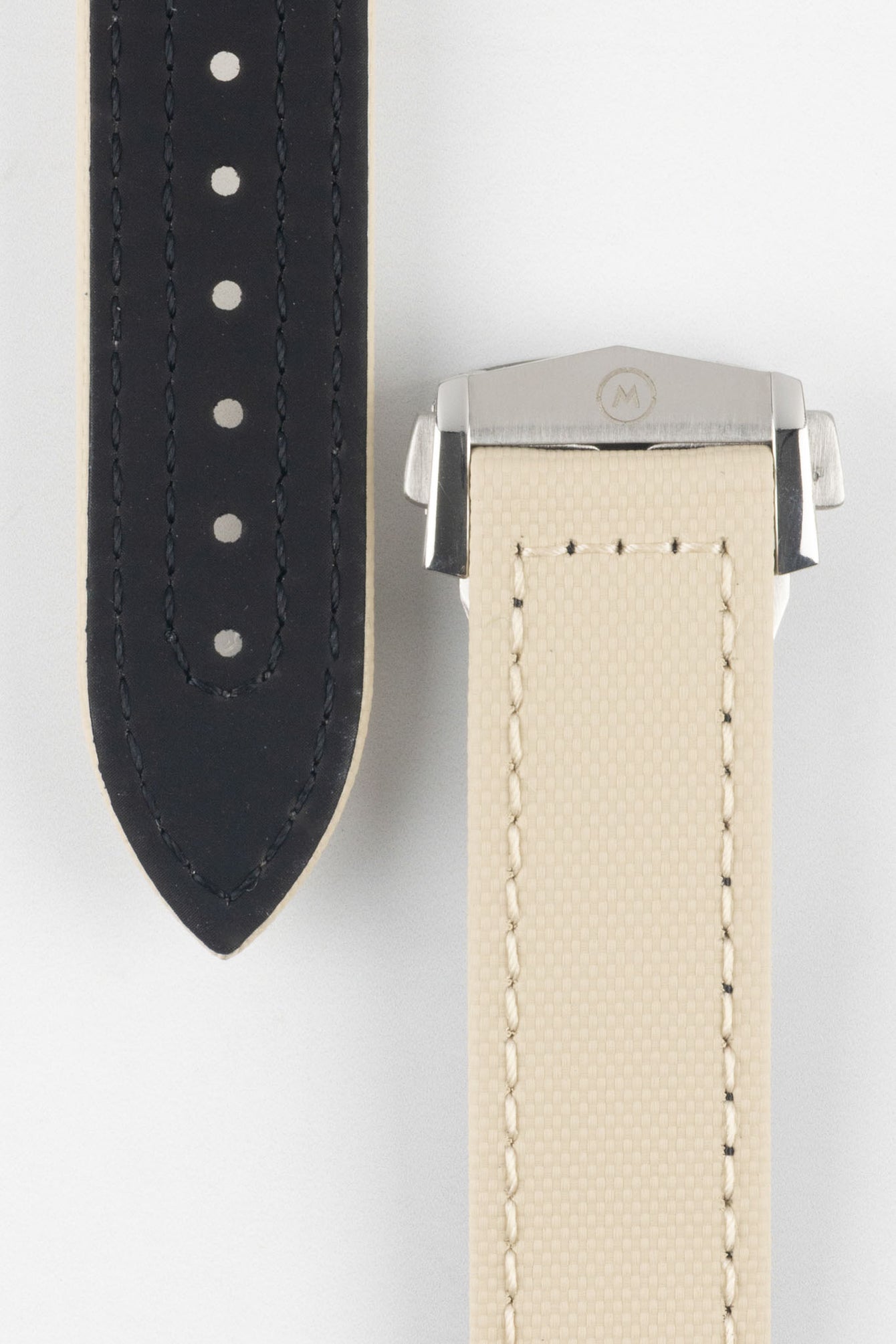 Close-up of a sand-colored textured watch strap with light stitching, showing one section with evenly spaced adjustment holes and the other attached to a brushed silver deployant clasp with an engraved logo, set against a light grey background.