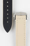 Close-up of a sand-colored textured watch strap with light stitching, showing one section with evenly spaced adjustment holes and the other attached to a brushed silver deployant clasp with an engraved logo, set against a light grey background.