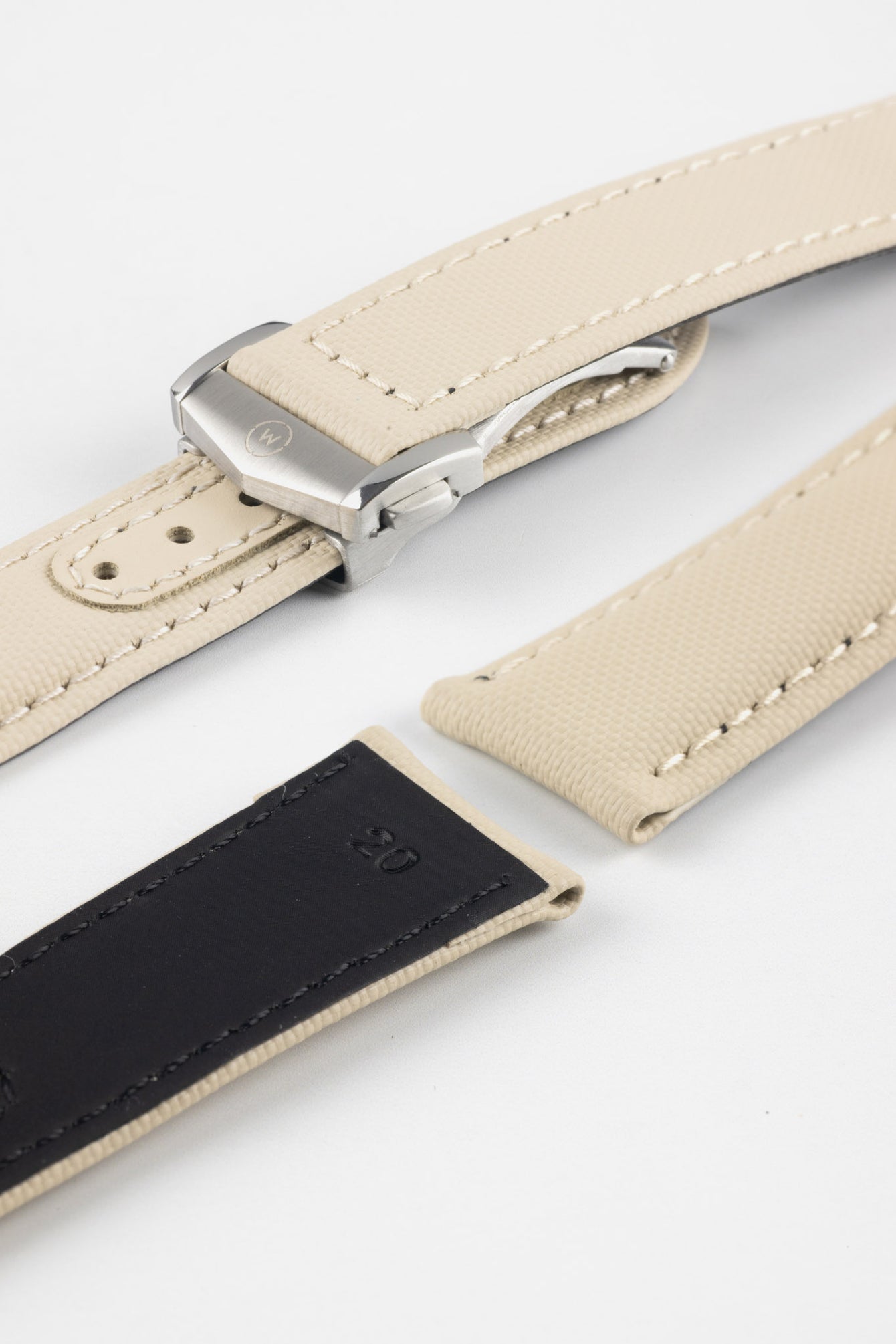 Angled close-up of a sand-colored textured watch strap with light stitching, featuring a brushed silver deployant clasp with an engraved logo, alongside separate strap sections, all arranged on a light grey background.