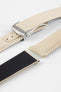 Angled close-up of a sand-colored textured watch strap with light stitching, featuring a brushed silver deployant clasp with an engraved logo, alongside separate strap sections, all arranged on a light grey background.