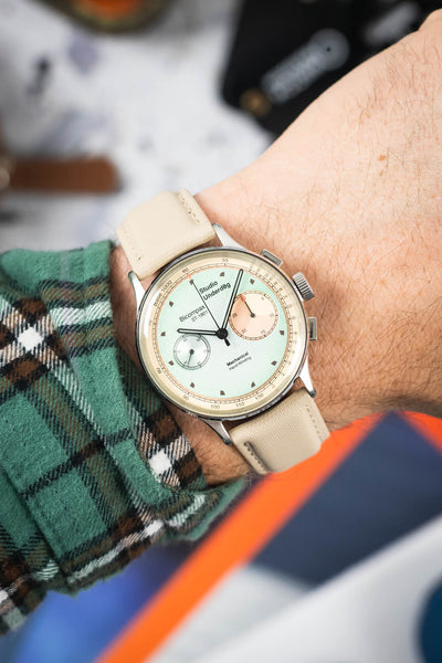 Close-up of a Studio Underd0g bicompax chronograph with a pastel green dial and subdials, worn on a wrist with a sand-colored textured strap. The polished case and pushers are visible, while a green plaid sleeve and blurred background add contrast and depth.
