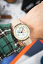Close-up of a Studio Underd0g bicompax chronograph with a pastel green dial and subdials, worn on a wrist with a sand-colored textured strap. The polished case and pushers are visible, while a green plaid sleeve and blurred background add contrast and depth.