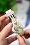 Close-up of a sand-colored textured watch strap with a stainless steel deployant clasp, held in hands to display the folding mechanism. The inner lining and adjustment holes are visible, with fine stitching details along the strap, set against a softly blurred background with hints of green and neutral tones.