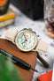 Close-up of a Studio Underd0g bicompax chronograph with a pastel green dial and cream subdials, resting on a brown leather pouch. The watch is paired with a sand-colored textured strap, while a blurred background of tools, a drink, and workshop-style elements adds depth and warm contrast.