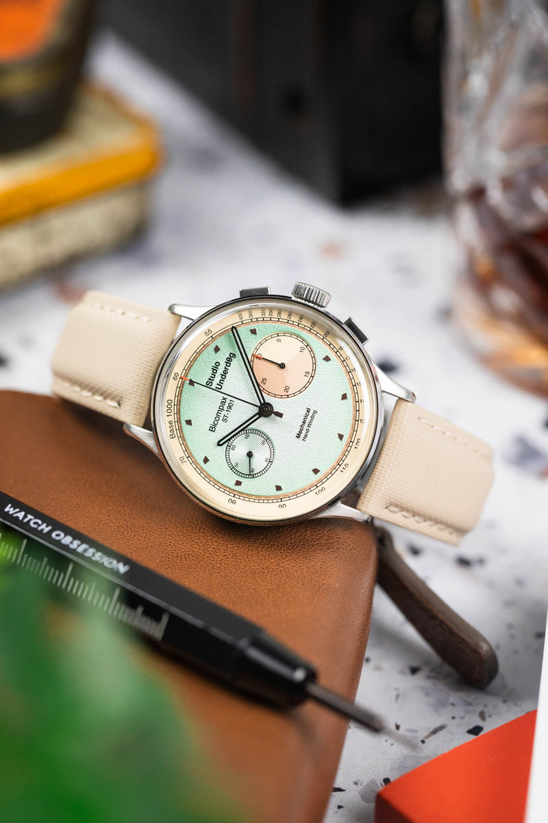 Close-up of a Studio Underd0g bicompax chronograph with a pastel green dial and cream subdials, resting on a brown leather pouch. The watch is paired with a sand-colored textured strap, while a blurred background of tools, a drink, and workshop-style elements adds depth and warm contrast.