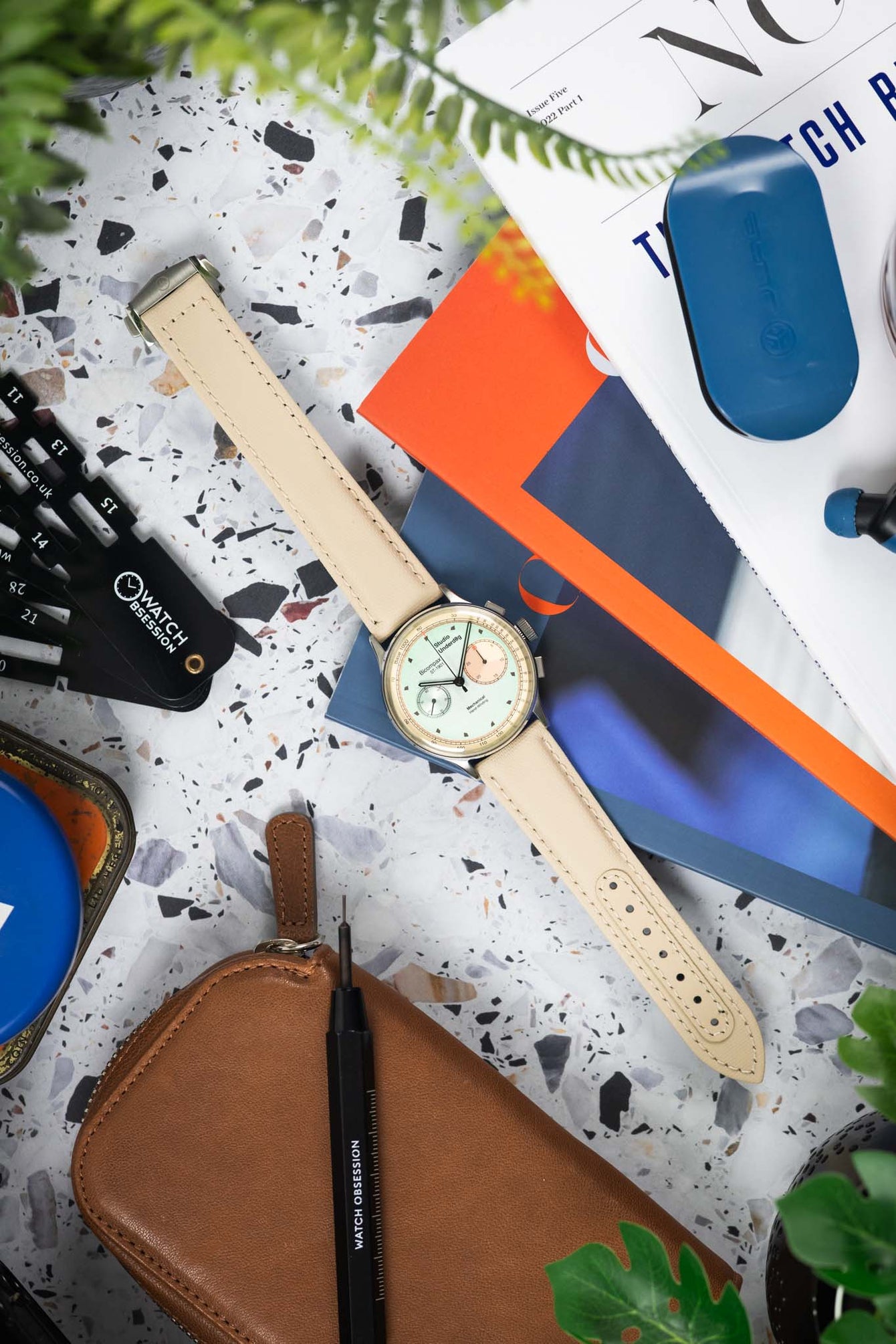 Flat lay of a Studio Underd0g bicompax chronograph with a pastel green dial and cream subdials on a sand-colored textured strap, arranged on a terrazzo-style surface. The scene includes a brown leather pouch, watch tools, magazines, earbuds, and greenery, creating a colorful workshop-style composition with orange, blue, and neutral accents.