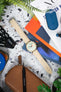 Flat lay of a Studio Underd0g bicompax chronograph with a pastel green dial and cream subdials on a sand-colored textured strap, arranged on a terrazzo-style surface. The scene includes a brown leather pouch, watch tools, magazines, earbuds, and greenery, creating a colorful workshop-style composition with orange, blue, and neutral accents.