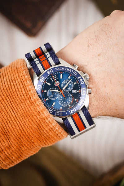 Premium TAG Heuer Formula 1 Gulf Edition chronograph watch featuring a stainless steel case, blue sunburst dial, three chronograph subdials, date display, blue tachymeter bezel, and a blue, white, and orange striped NATO-style fabric strap. Wrist-shot lifestyle photograph showcasing iconic Gulf racing colours, Swiss craftsmanship, precision chronograph performance, exceptional comfort, and bold motorsport-inspired styling for collectors, enthusiasts, and everyday wear.