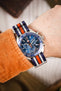 Premium TAG Heuer Formula 1 Gulf Edition chronograph watch featuring a stainless steel case, blue sunburst dial, three chronograph subdials, date display, blue tachymeter bezel, and a blue, white, and orange striped NATO-style fabric strap. Wrist-shot lifestyle photograph showcasing iconic Gulf racing colours, Swiss craftsmanship, precision chronograph performance, exceptional comfort, and bold motorsport-inspired styling for collectors, enthusiasts, and everyday wear.