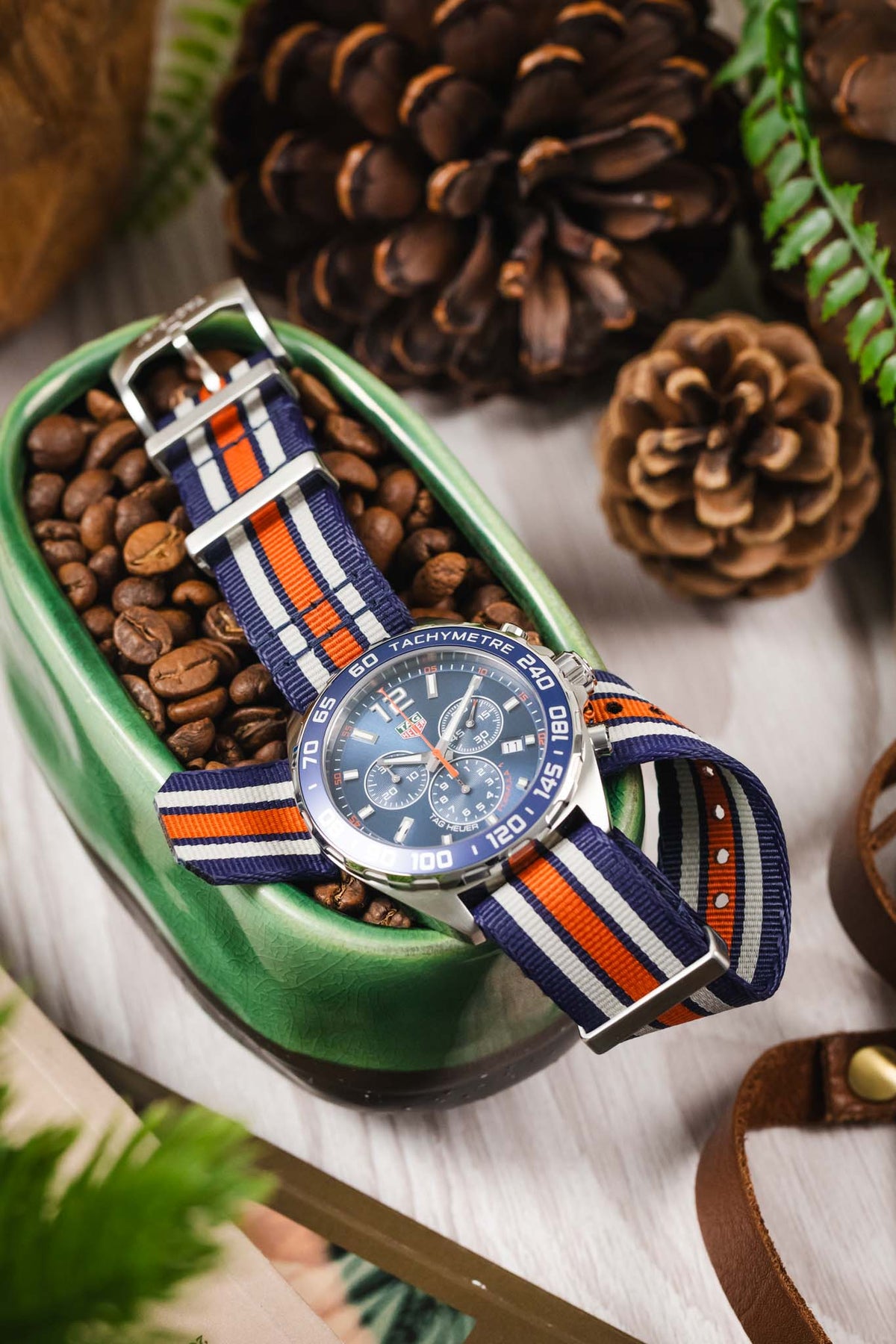 TAG Heuer Formula 1 Gulf Edition chronograph with a blue, white, and orange NATO strap displayed in a dish of coffee beans with lifestyle accessories.