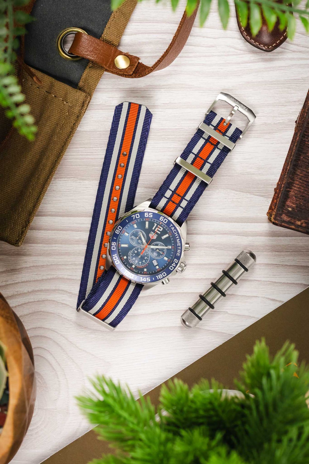 Premium TAG Heuer Formula 1 Gulf Edition chronograph watch featuring a stainless steel case, blue sunburst dial, chronograph subdials, date display, blue tachymeter bezel, and a blue, white, and orange striped NATO-style fabric strap. Lifestyle flat-lay photograph showcasing iconic Gulf racing colours, Swiss craftsmanship, sporty chronograph performance, and distinctive motorsport-inspired styling alongside watchmaking tools, leather accessories, and contemporary lifestyle décor.