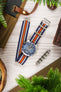 Premium TAG Heuer Formula 1 Gulf Edition chronograph watch featuring a stainless steel case, blue sunburst dial, chronograph subdials, date display, blue tachymeter bezel, and a blue, white, and orange striped NATO-style fabric strap. Lifestyle flat-lay photograph showcasing iconic Gulf racing colours, Swiss craftsmanship, sporty chronograph performance, and distinctive motorsport-inspired styling alongside watchmaking tools, leather accessories, and contemporary lifestyle décor.