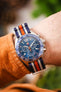Premium TAG Heuer Formula 1 Gulf Edition chronograph watch featuring a stainless steel case, blue sunburst dial, chronograph subdials, date display, blue tachymeter bezel, and a blue, white, and orange striped NATO-style fabric strap. Wrist-shot lifestyle photograph showcasing iconic Gulf racing colours, Swiss craftsmanship, sporty chronograph performance, exceptional comfort, and bold motorsport-inspired styling for collectors, enthusiasts, and everyday wear.