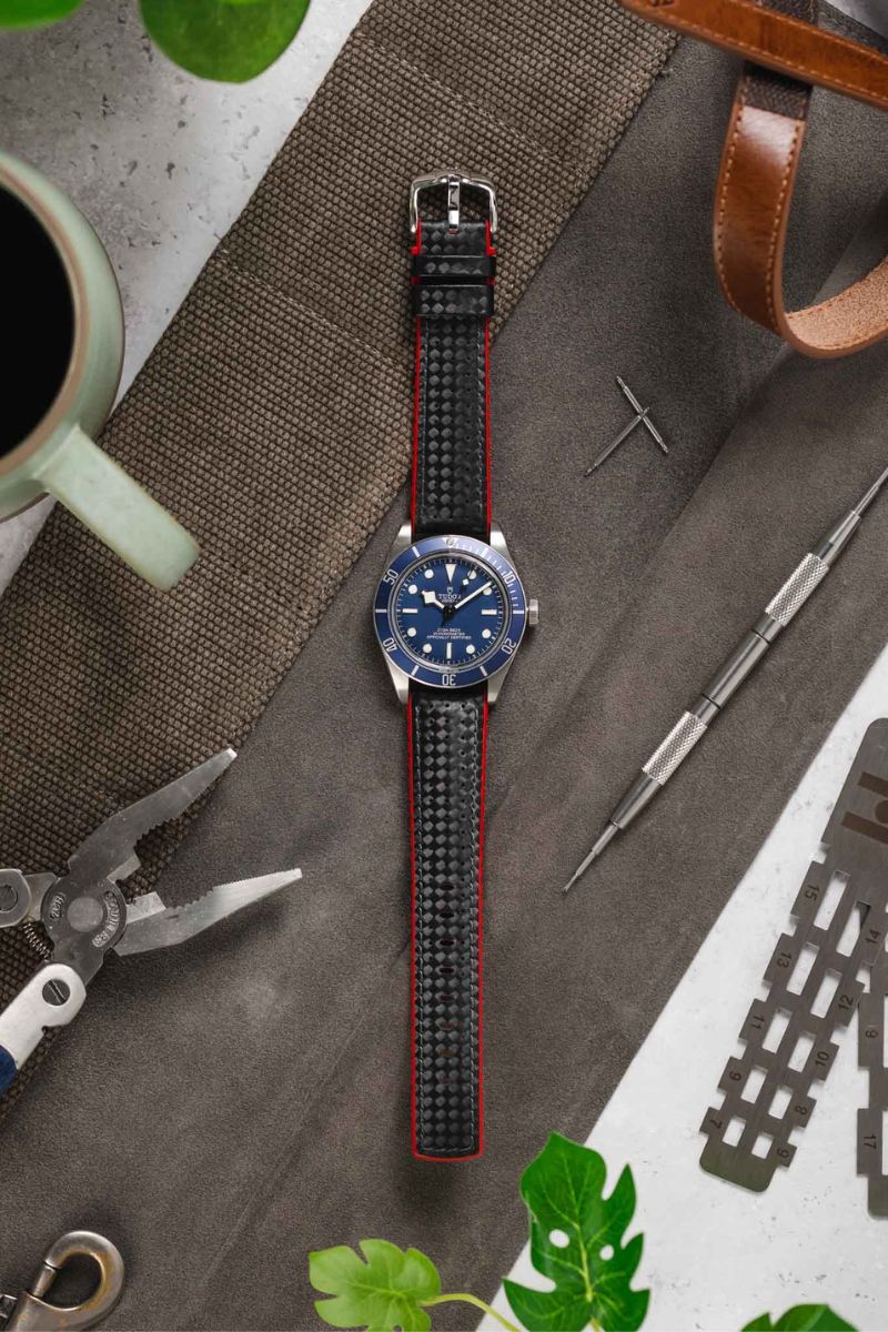 Flat lay of a stainless steel dive watch with a blue dial attached to a black textured strap with red edges, arranged on a work surface. Surrounding items include a watchmaker’s tool, calipers, pliers, a coffee mug, leather strap, and green leaves, all set against a mix of fabric and light grey background.