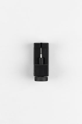 A black watch bracelet link removal tool is positioned from a top-down perspective against a plain white background. The tool has a small pin in the center with a ridged screw on the bottom, used to push out pins from watch bracelet links.