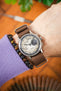 Close-up of a Studio Underd0g Bicompax watch with a cream dual-subdial dial and brown ribbed nylon strap, worn on a wrist alongside a beaded bracelet, against a softly blurred warm-toned background.