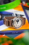 Close-up of a Studio Underd0g Bicompax chronograph watch with a textured cream dial and dual subdials, paired with a brown ribbed nylon strap. The watch rests on stacked books (one labeled “ISSUE .88”) beside a blurred green headphone band, with a colorful background of orange, blue, and green tones.
