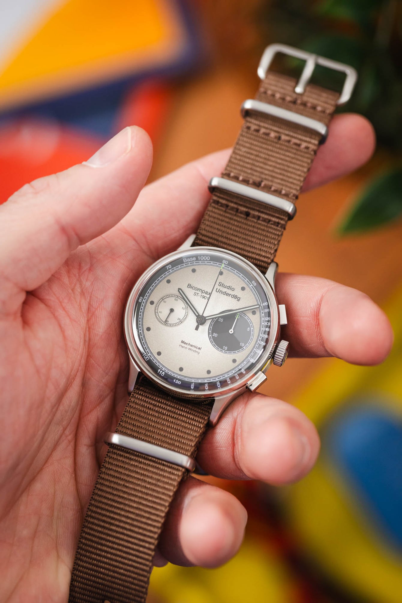 Close-up of a Studio Underd0g Bicompax watch with a textured cream dial and two subdials, held in a hand and paired with a brown ribbed nylon strap. The brushed metal case and NATO-style strap hardware are in sharp focus, with a softly blurred, colorful background of orange, green, and blue tones.