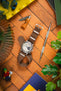 Flat-lay of a Studio Underd0g Bicompax watch with a cream dual-subdial face and brown ribbed nylon strap on a wooden surface, surrounded by a small folding knife, precision screwdriver, earbuds in a yellow tray, green plant leaves, a toy figure, and colorful books.