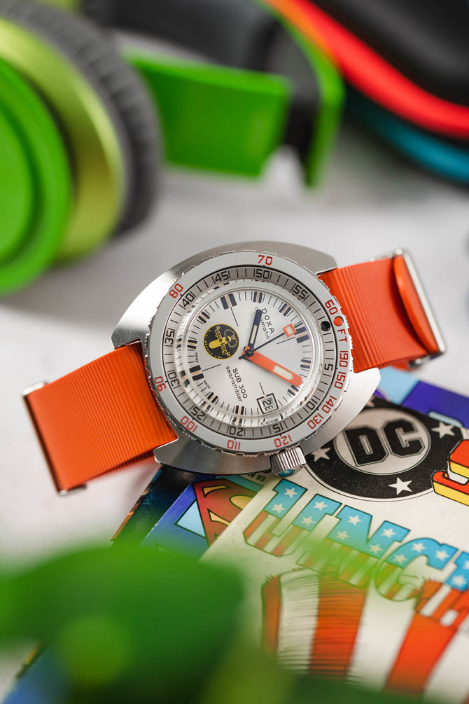 Doxa sub 300 searambler silver lung limited edition fitted with orange bonetto cinturini 328 premium rubber one piece watch strap curved on top of comic books on table