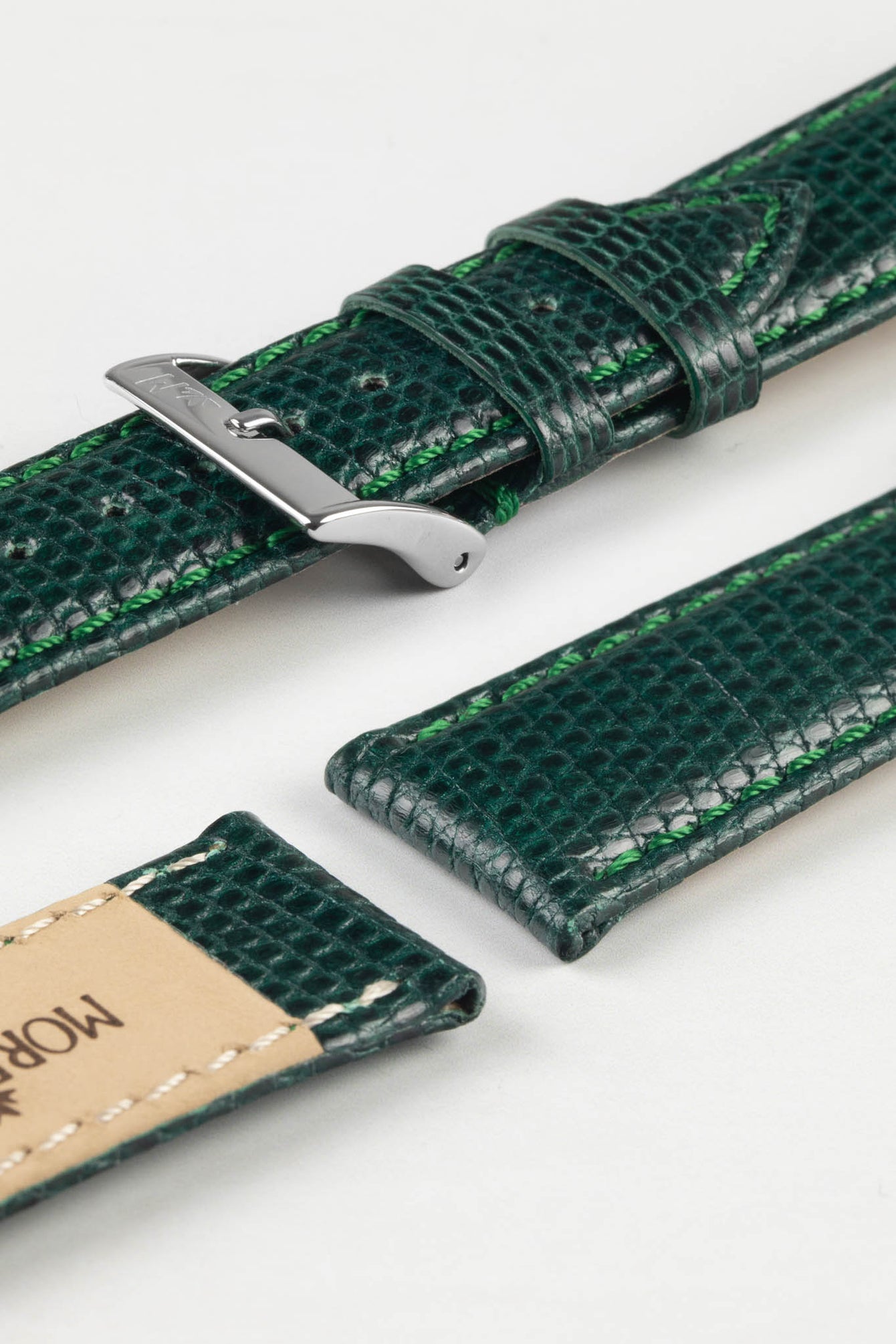 A close-up, top-down shot of a dark green watch strap with a lizard-skin texture, lying in two pieces on a white surface. The upper piece features a silver-toned buckle, while the lower piece shows the light brown leather underside with a partially visible "MORELLATO" stamp. Green stitching is visible along the edges of the strap.